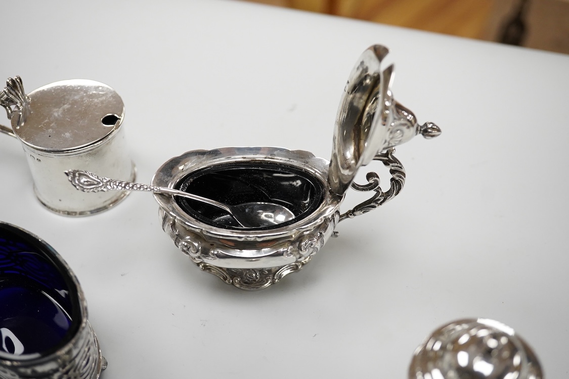 An ornate Edwardian silver oval mustard pot and matching spoon by Barker Brothers, Birmingham, 1902, length, 95mm, together with five other assorted silver condiments including a pair of pierced oval salts with blue glas